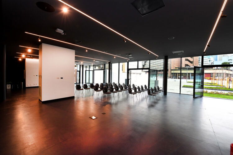 Sala eventi con sedie disposte a platea, luminosa con grandi vetrate che affacciano su uno spazio esterno.