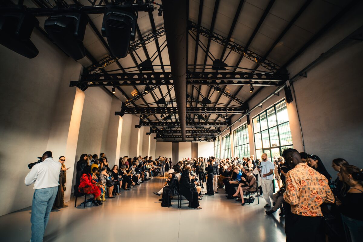**Alt text:** Spazio industriale con soffitto a capriate pieno di persone sedute per una sfilata di moda.