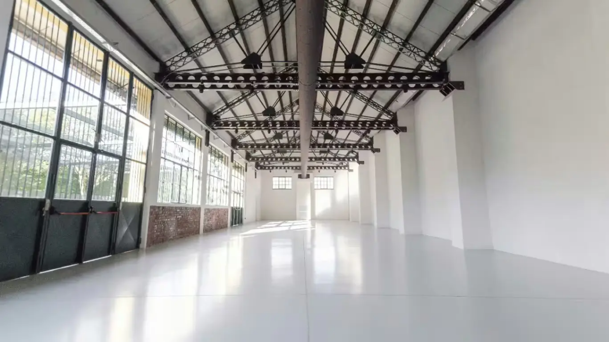 **Alt text:** Ampio spazio industriale vuoto con pavimento bianco, pareti chiare e soffitto con travi in metallo.