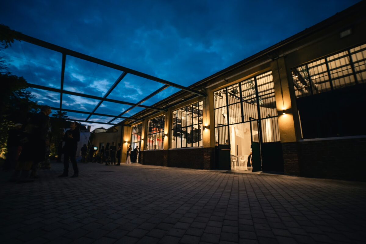 Esterno di un edificio industriale con ampie vetrate e illuminazione serale, persone in conversazione nel cortile.