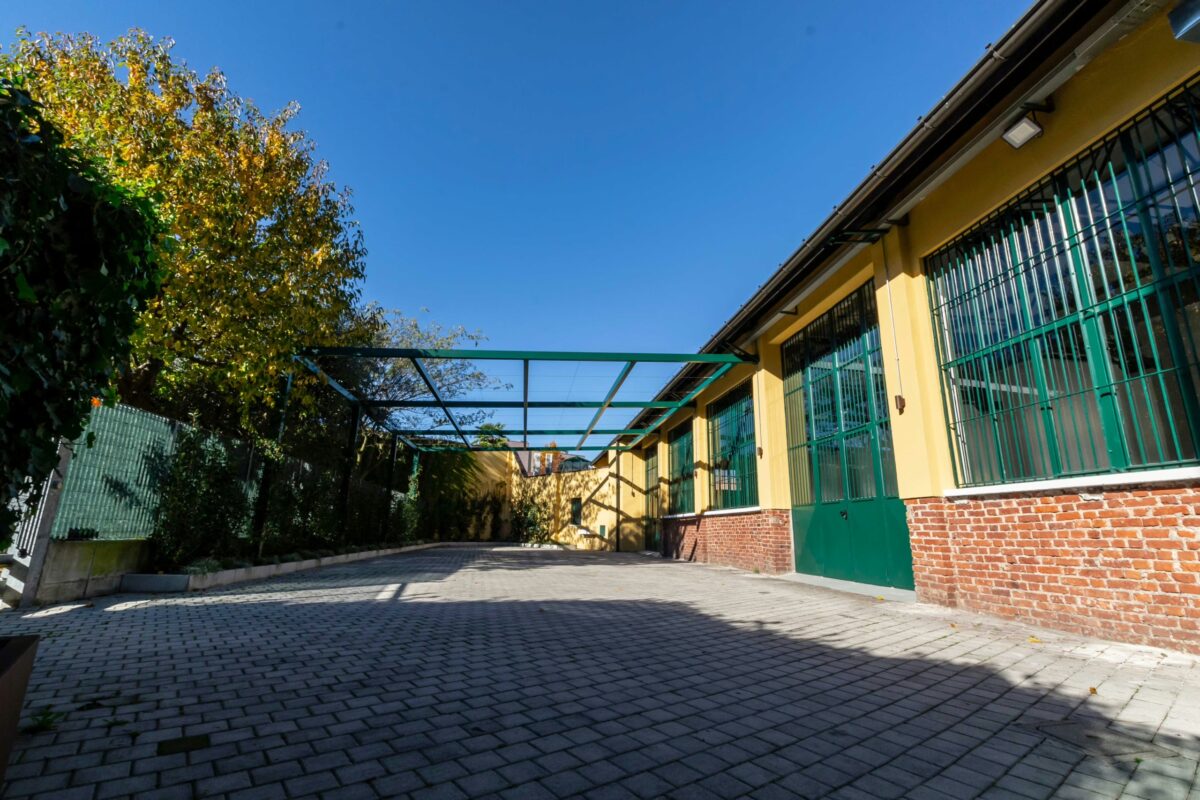 Vista diurna del cortile pavimentato con pergolato metallico e facciata con vetrate e pareti giallo ocra.