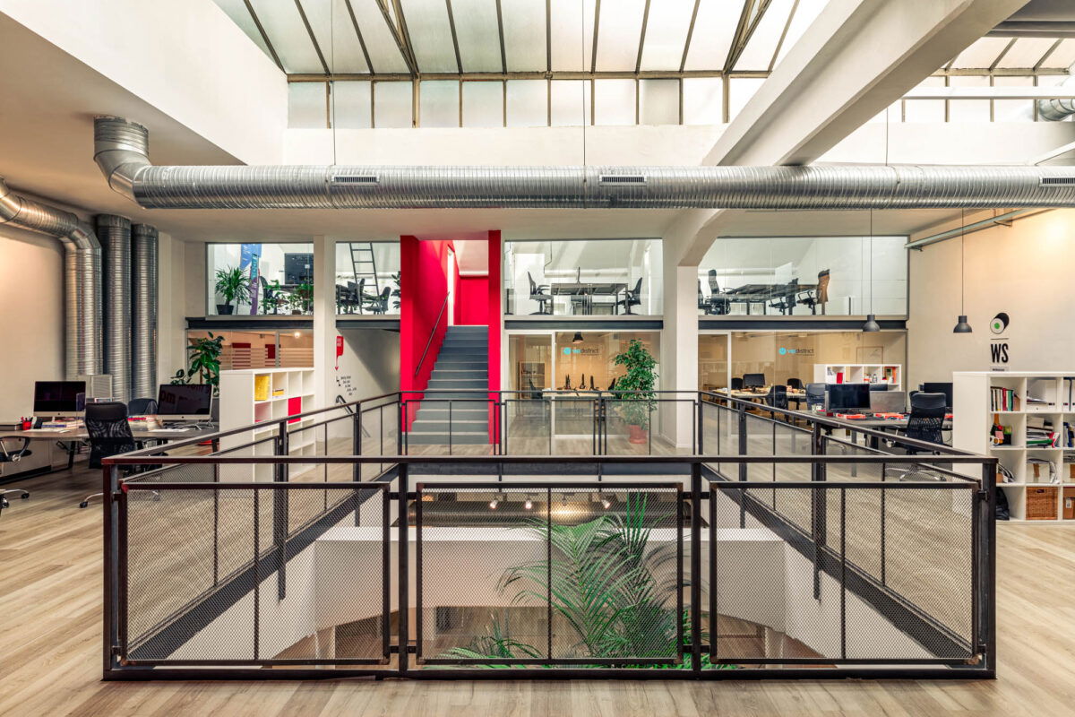 Area coworking su due livelli con grandi vetrate, pareti bianche e scala rossa centrale.