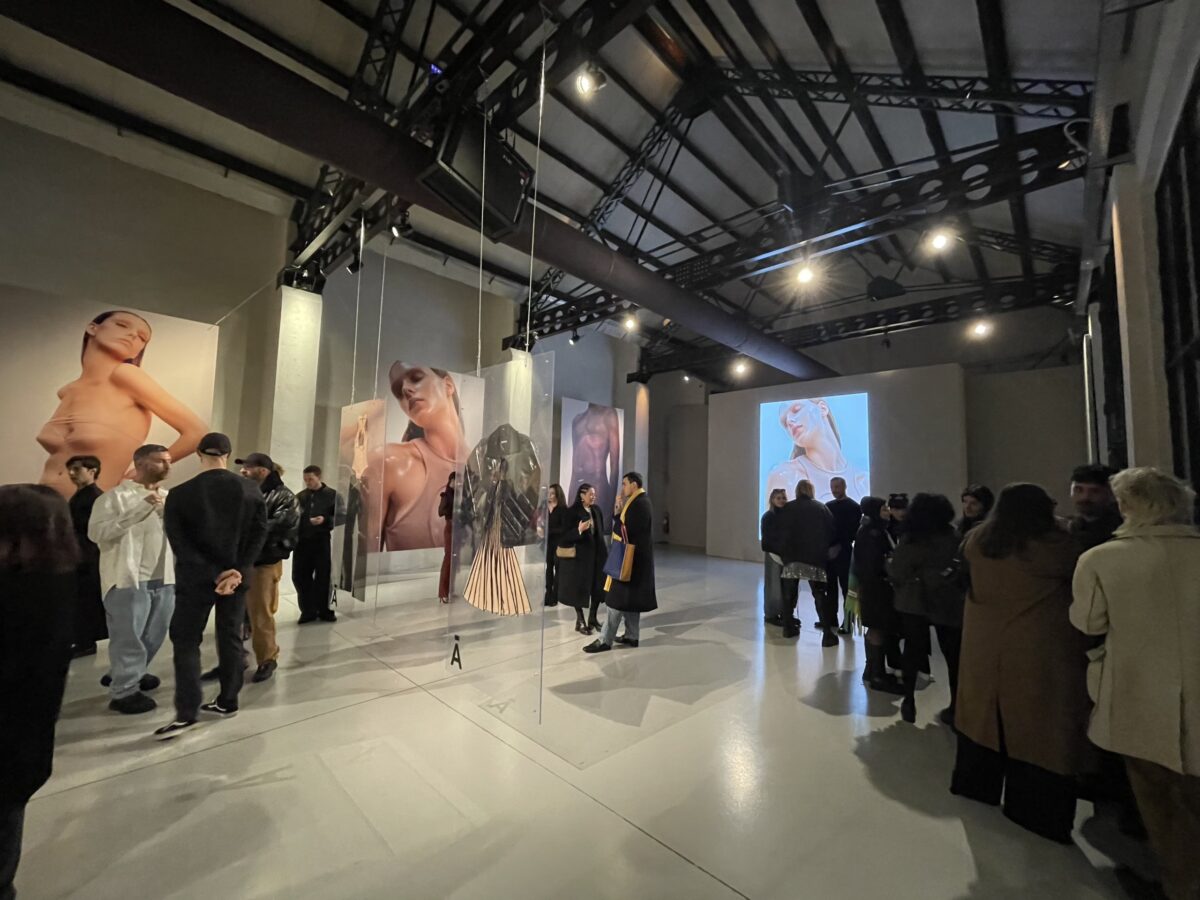 Interno industriale con soffitto in metallo e installazione fotografica, persone raccolte in piccoli gruppi durante una mostra.