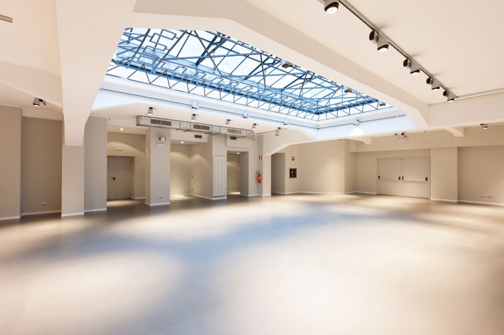 Sala moderna e minimalista con ampio lucernario, tavoli bianchi e piante verdi, ideale per meeting ed eventi aziendali.