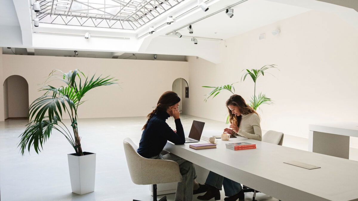Due donne lavorano a un tavolo bianco sotto un grande lucernario.